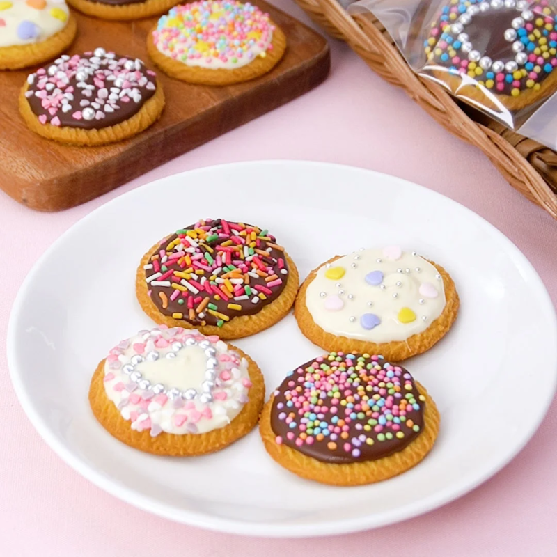 カラフルなスプリンクルやチョコレート、アイシングで可愛くデコレーションされた丸いクッキーが、白いお皿や木製のボードに複数並べられています。プレゼントにもぴったりの華やかなスイーツです。
