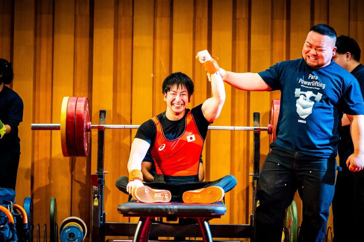 パワーリフティングの試合で、日本の選手がベンチプレスを成功させ、満面の笑みで喜びを表現している瞬間。隣の男性も笑顔で祝福しており、達成感と喜びが伝わる一枚です。