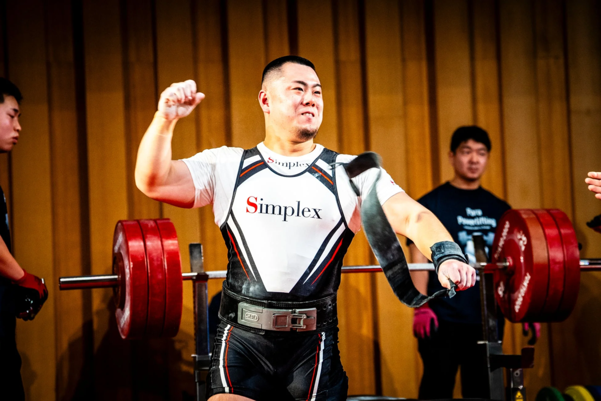 パワーリフティングの大会で、シングレットを着た男性が成功したリフトを祝って力強くポーズをとっている様子。背景にはバーベルと他の参加者が見える。