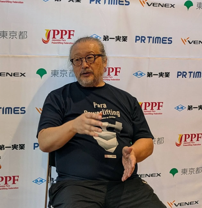 眼鏡をかけ、髭と長髪の男性が、パラスポーツイベントの背景を背に、パラパワーリフティングのTシャツを着て話している様子。スポンサーロゴが多数見られ、記者会見かインタビューの場面と推測される。