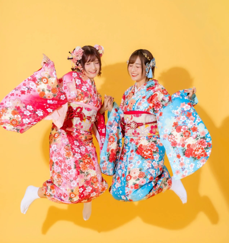 鮮やかな花柄の振袖を着た二人の女性が、手をつないで楽しそうにジャンプしている様子