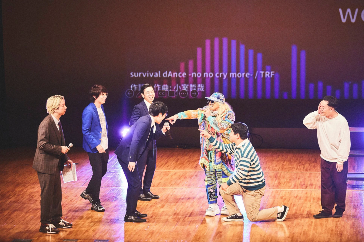 ステージ上で複数の男性がパフォーマンスやトークを行っており、背景のスクリーンにはTRFの楽曲「survival dAnce -no no cry more-」の情報が表示されている