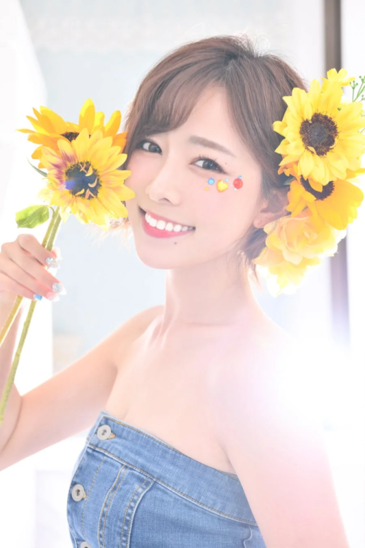 笑顔の女性がひまわりを手に持ち、肩出しのデニムトップスを着てポーズをとっている