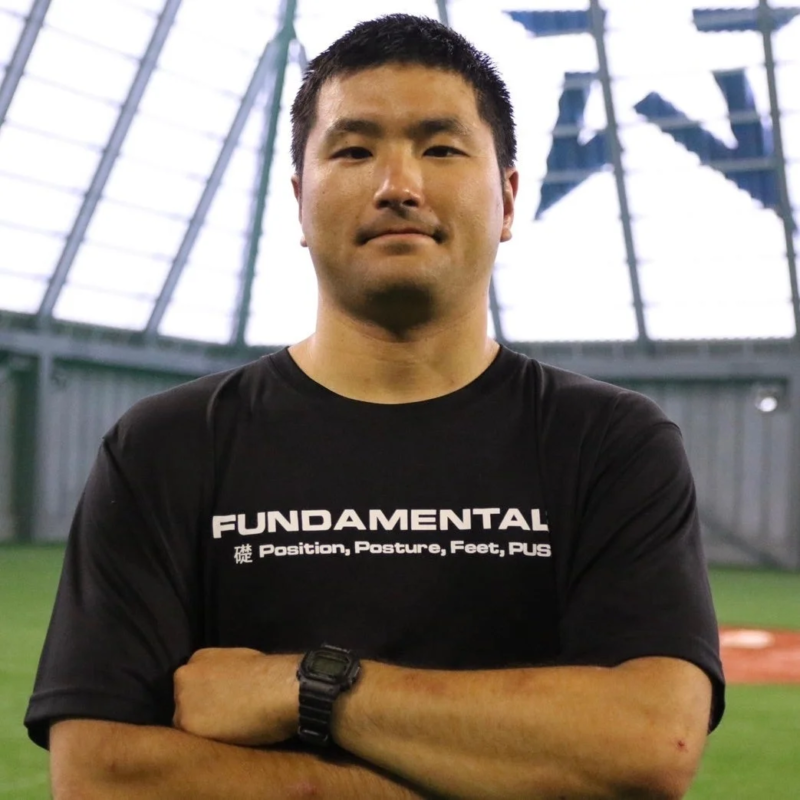 黒いTシャツを着たアジア人男性が腕を組み、カメラに向かって立っています。Tシャツには「FUNDAMENTAL」という文字が見えます。背景は屋内のスポーツ施設で、野球場のような緑色の地面と白いドーム型の天井が特徴です。