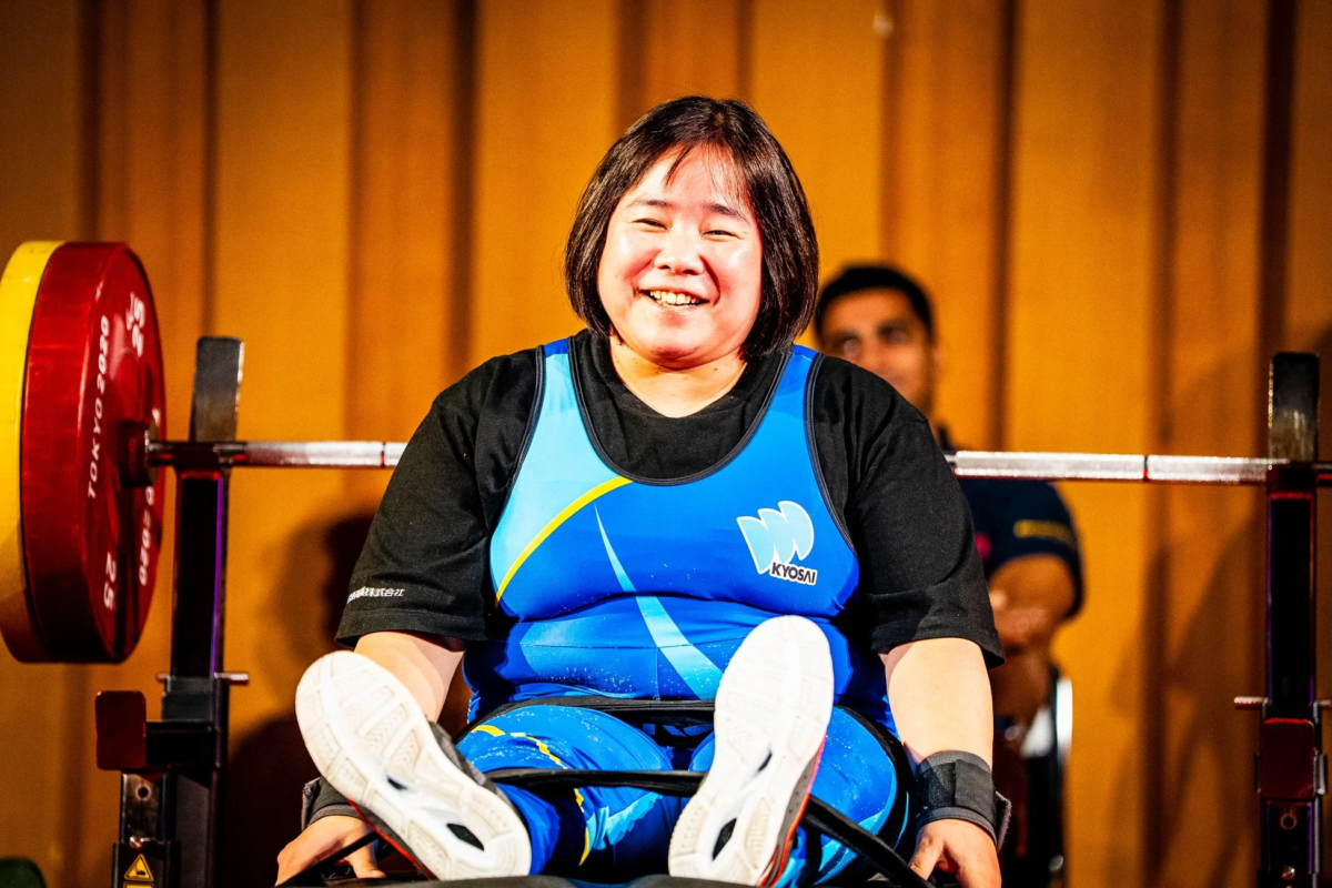 青いユニフォームを着た女性アスリートが、ベンチプレス台の前で満面の笑みを浮かべています。東京2020のロゴが入った重りがバーベルにセットされており、競技中の様子を捉えたものと思われます。