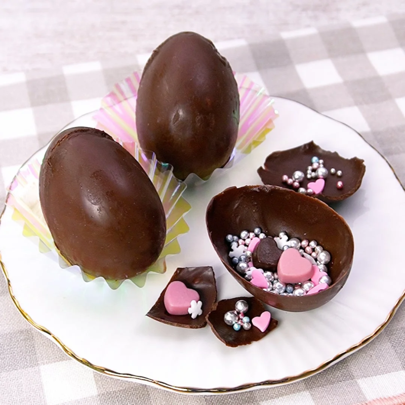 白い皿に盛られたチョコレート製のイースターエッグ。2つはそのままの形で、1つは割れており、中には銀色の粒やピンクのハート型チョコレートが詰まっています。可愛らしい手作りスイーツのようです。