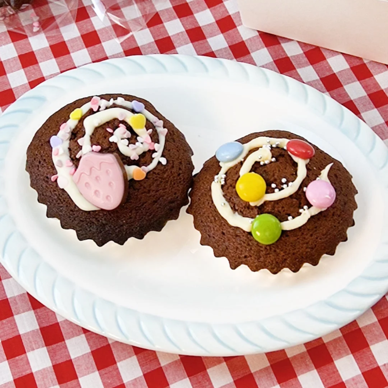 赤と白のチェック柄のテーブルクロスの上に置かれた白い皿に、デコレーションされたチョコレートカップケーキが2つ並んでいます。片方にはイチゴの飾り、もう片方にはカラフルなチョコが載っています。