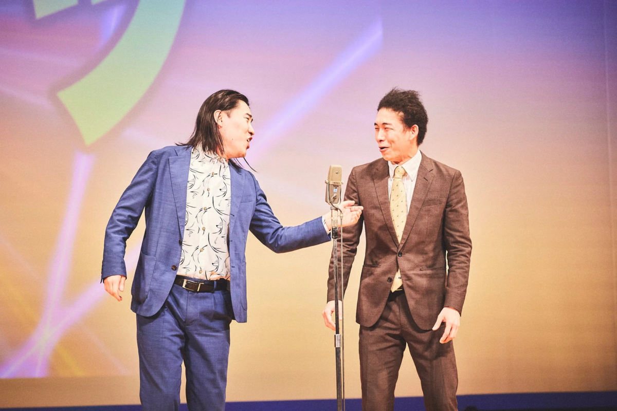 舞台上で二人の男性がマイクを挟んで対話している様子