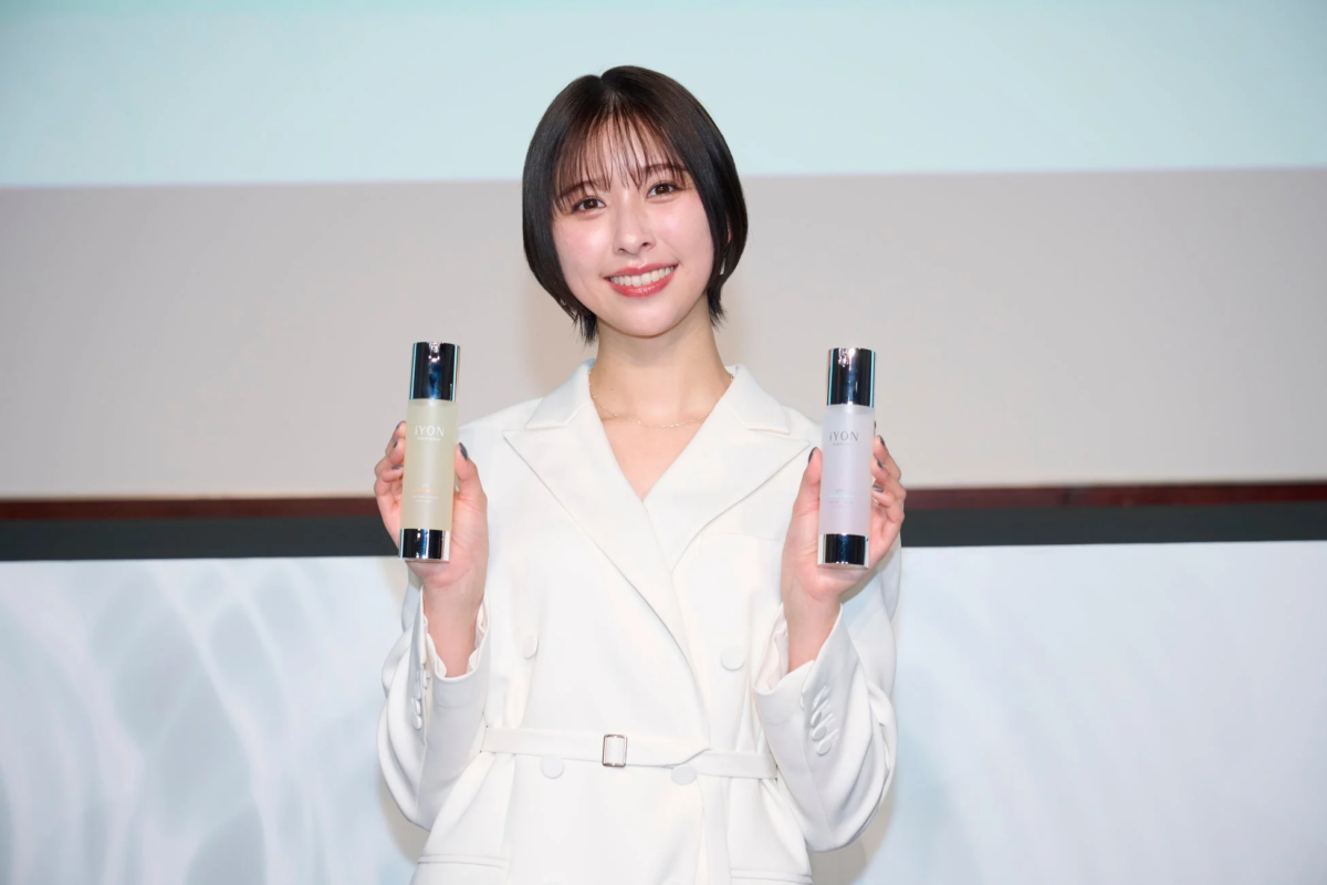白いジャケットを着た若い日本人女性が、笑顔で2本の「iYON」スキンケア製品のボトルを両手に持って見せている画像です。製品発表会のような雰囲気です。