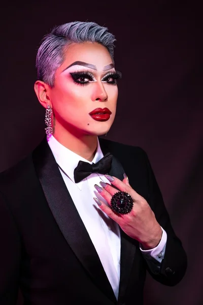 鮮やかなドラァグメイクを施した人物が、黒いタキシードと蝶ネクタイを着用しポーズをとっています。銀色の髪、強調された目元、赤い口紅が特徴的で、自信に満ちた表情と華やかなアクセサリーが印象的です。