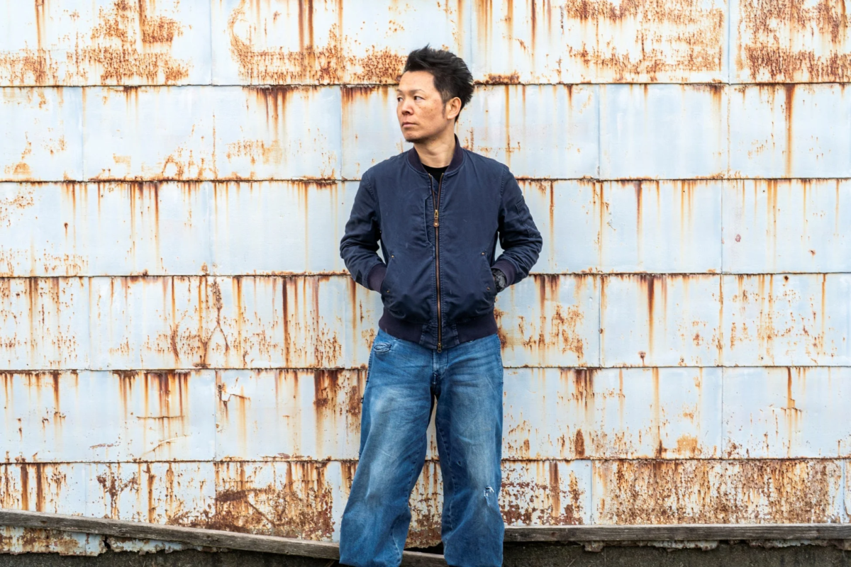 錆びた金属の壁を背景に、ブルゾンとデニムを着用した歌代隼人さん