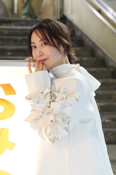 白い装飾的な袖のドレスを着た女性が、穏やかな笑顔でカメラを見つめています。背景には階段が見える屋内のポートレートです。