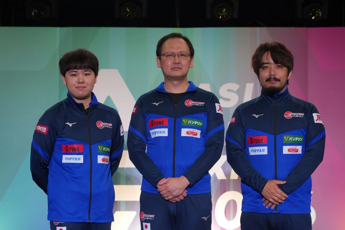 青いジャケットを着た3人の男性がステージに立っており、ジャケットにはESPORTS JAPAN、G TUNE、TOPPAN、バンテリンなどのロゴが見えます。eスポーツ関連のイベントか記者会見の様子です。