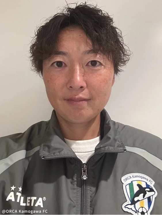 ORCA Kamogawa FCのエンブレムとATLETAのロゴが入ったグレーのジャケットを着用した女性のサッカー選手のポートレートです。短くウェーブのかかった髪型で、カメラをまっすぐ見ています。