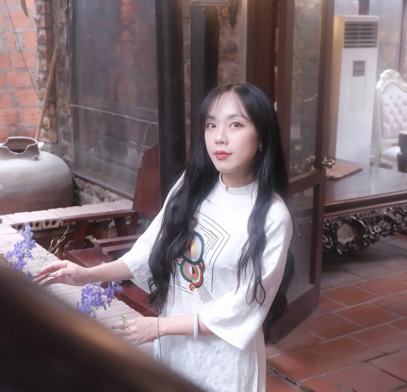 白いアオザイを着用した若い女性が、伝統的な内装の空間で、紫色の花に囲まれながら優雅にポーズをとっています