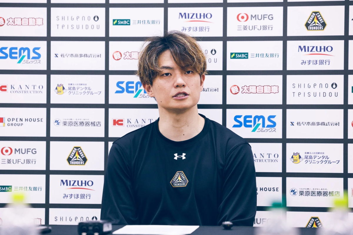 記者会見のような場所で、黒い長袖シャツを着た男性がカメラに向かって座っている