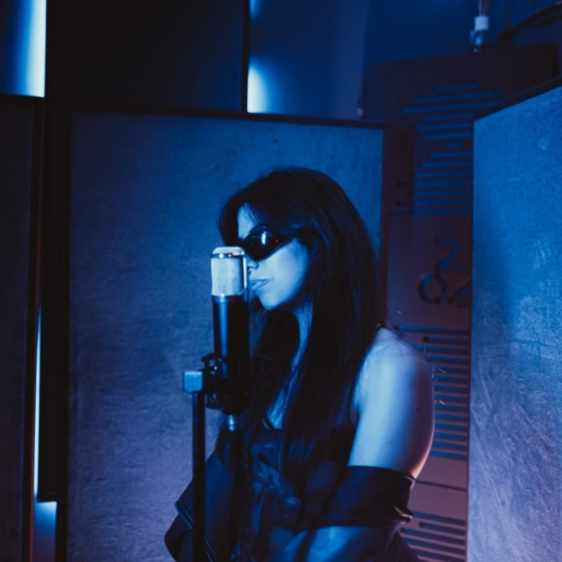 青い照明が印象的なレコーディングスタジオで、サングラスをかけた女性がマイクの前に立っています