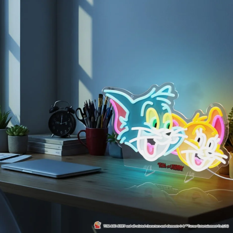 トムとジェリーのカラフルなネオンサインが特徴的なデスクの風景。木製の机にはノートパソコン、目覚まし時計、植物、文房具が整然と置かれ、アニメキャラクターのライトが部屋を明るく照らしています。落ち着いた雰囲気のインテリアが魅力的です。