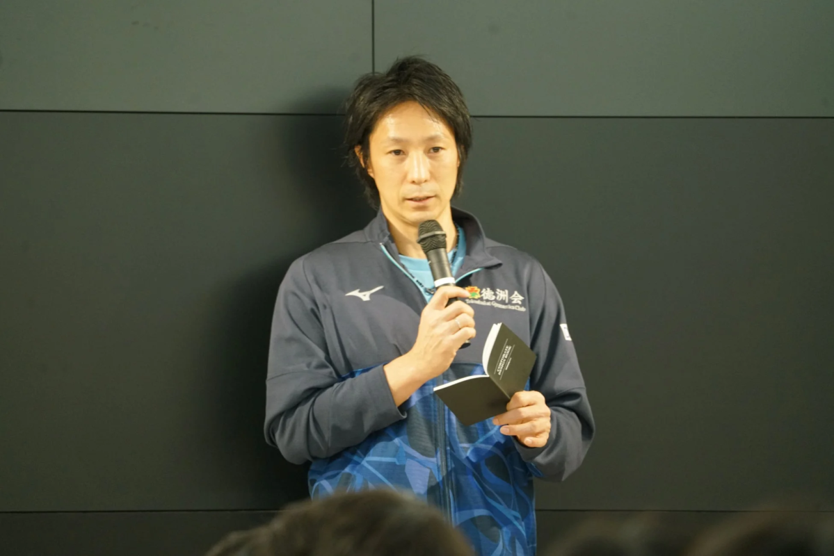 スポーツジャケットを着た男性がマイクを持ち、小さな本を片手に話している様子