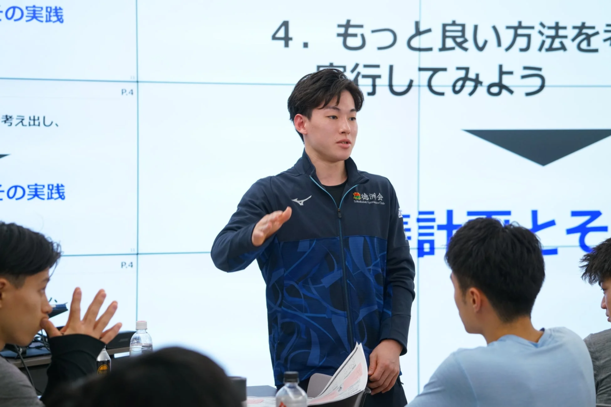 青いジャケットを着た男性が、スクリーンに映し出された文字の前で、聴衆に何かを説明している場面