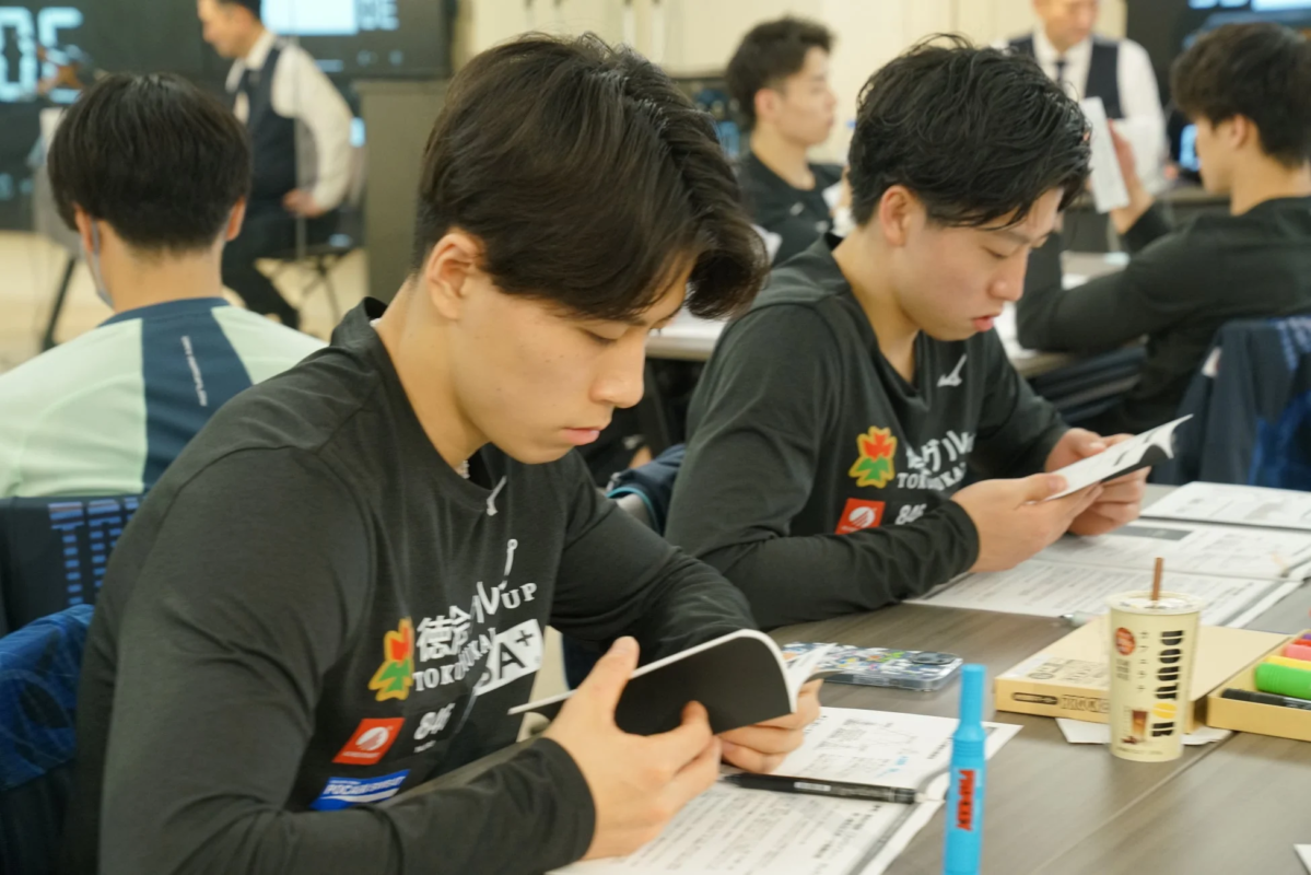 室内で数人の若い男性がテーブルで書類やノートを読み、集中している様子