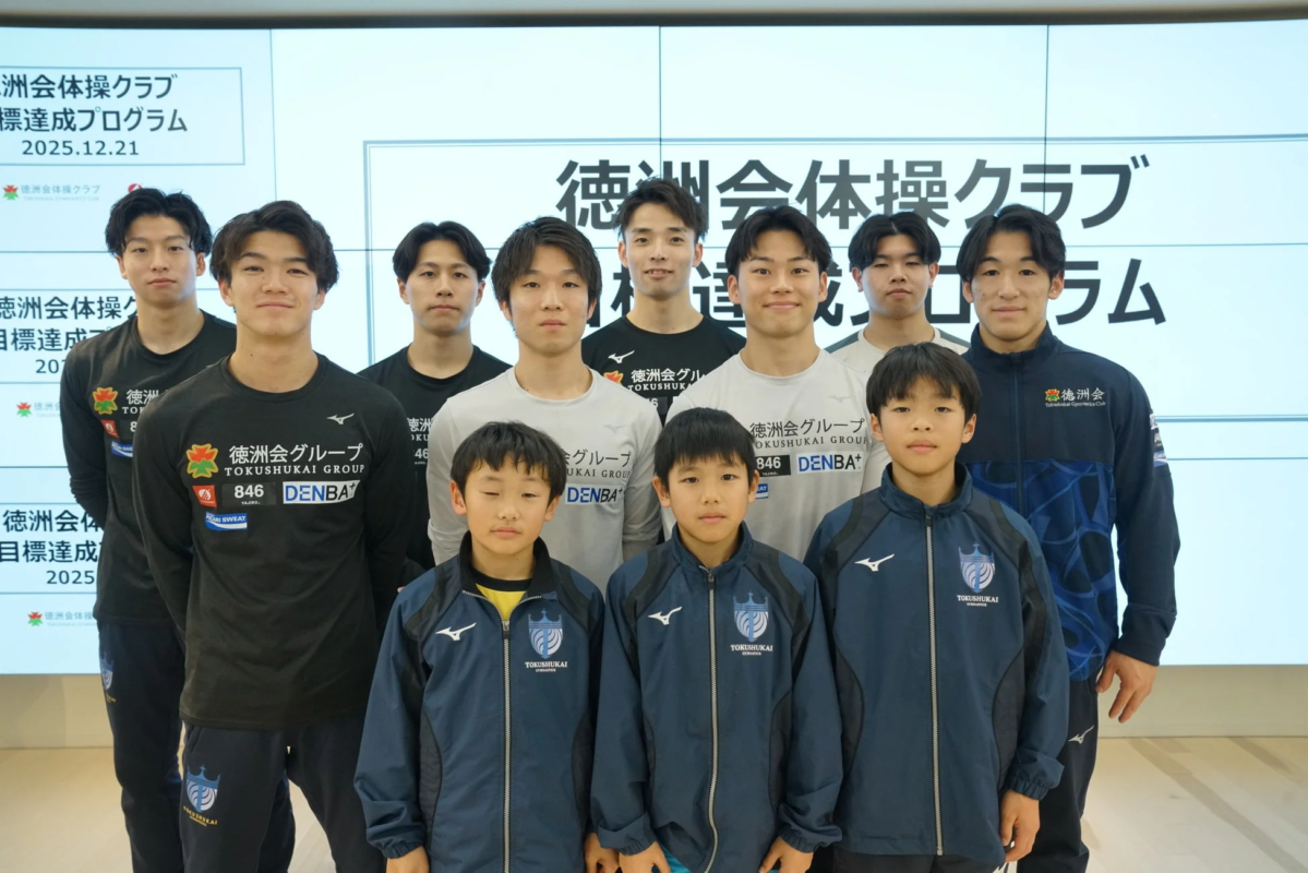徳洲会体操クラブのメンバーと思われる男性と子供たちが、クラブ名やスポンサーロゴが入ったウェアを着用し、目標達成プログラムが映し出されたスクリーンを背景に集合写真を撮っている様子
