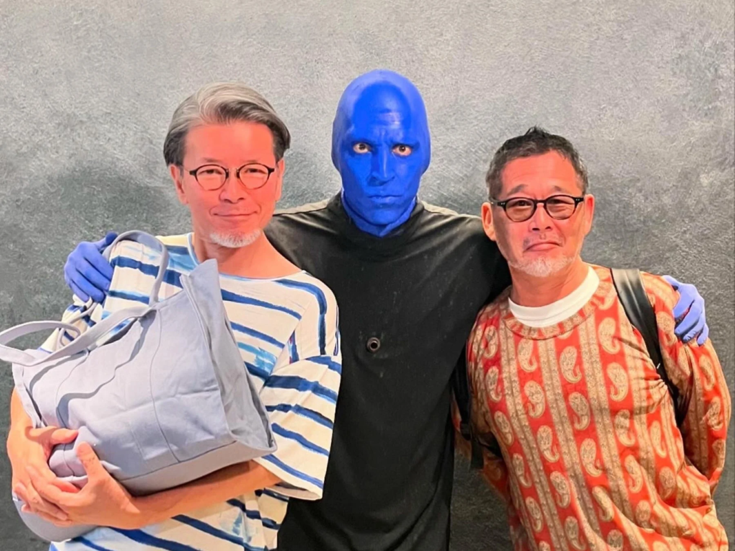 青い顔と手袋の人物が、眼鏡をかけた2人の男性と肩を組み、笑顔で並んで写っています。ブルーマングループのメンバーとファンまたは友人との記念撮影のようです。