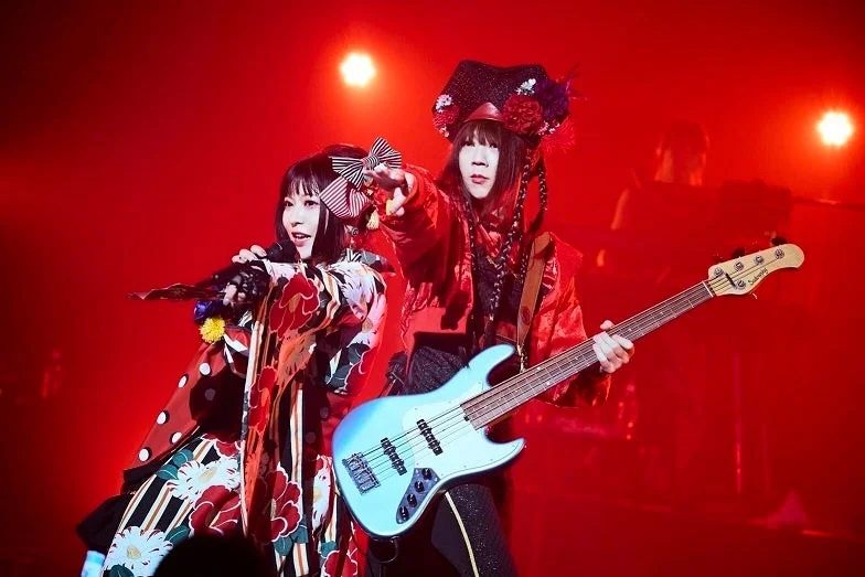赤い照明に照らされたステージで、和風の衣装をまとった女性ボーカルと、ベースを演奏するもう一人の女性がパフォーマンスをしている様子
