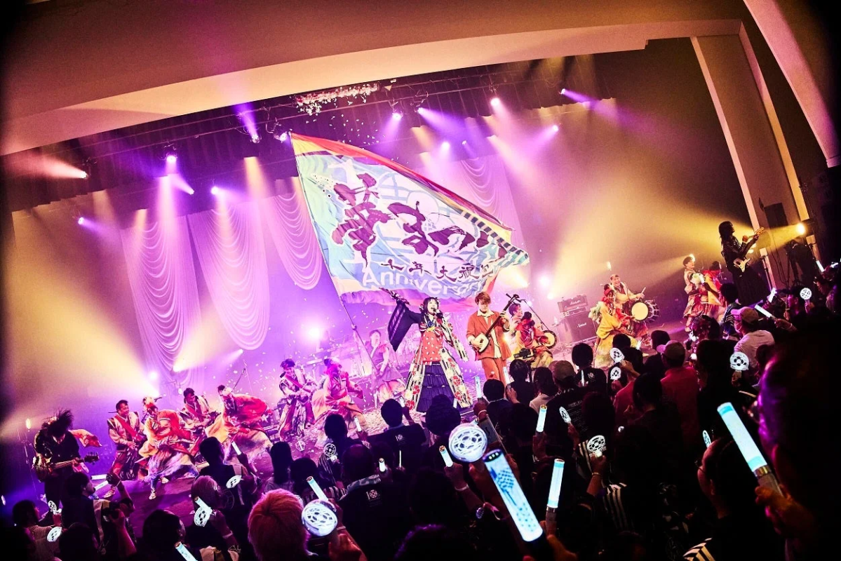 和楽器バンドのライブパフォーマンスを捉えた画像。ステージ上ではメンバーが和楽器を含む様々な楽器を演奏し、大きな旗が掲げられています。観客はペンライトを振って熱狂しており、紫色の照明が会場を彩っています。