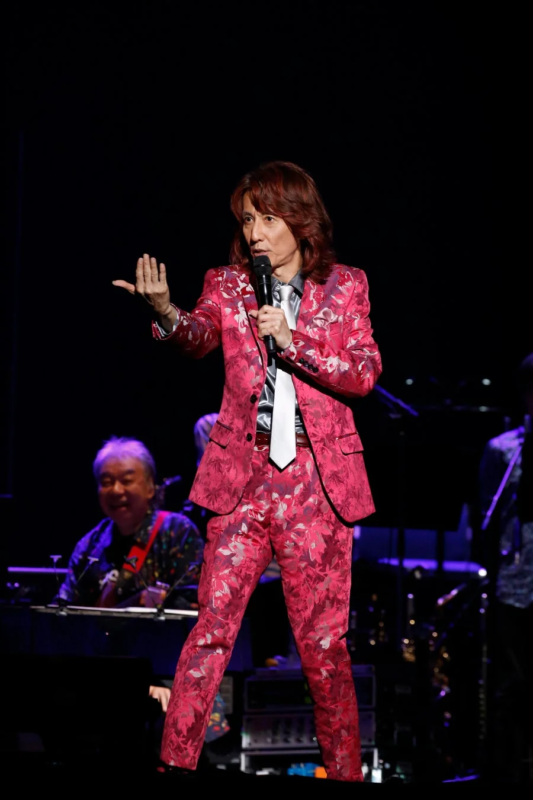 ピンクの花柄スーツを着た男性がステージで歌っている