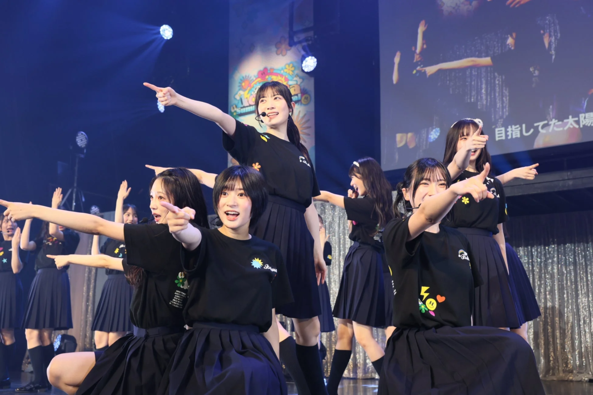 ステージ上で黒いTシャツとスカートを着用した女性アイドルグループが、笑顔で指を差すポーズを取りながらパフォーマンスしている様子