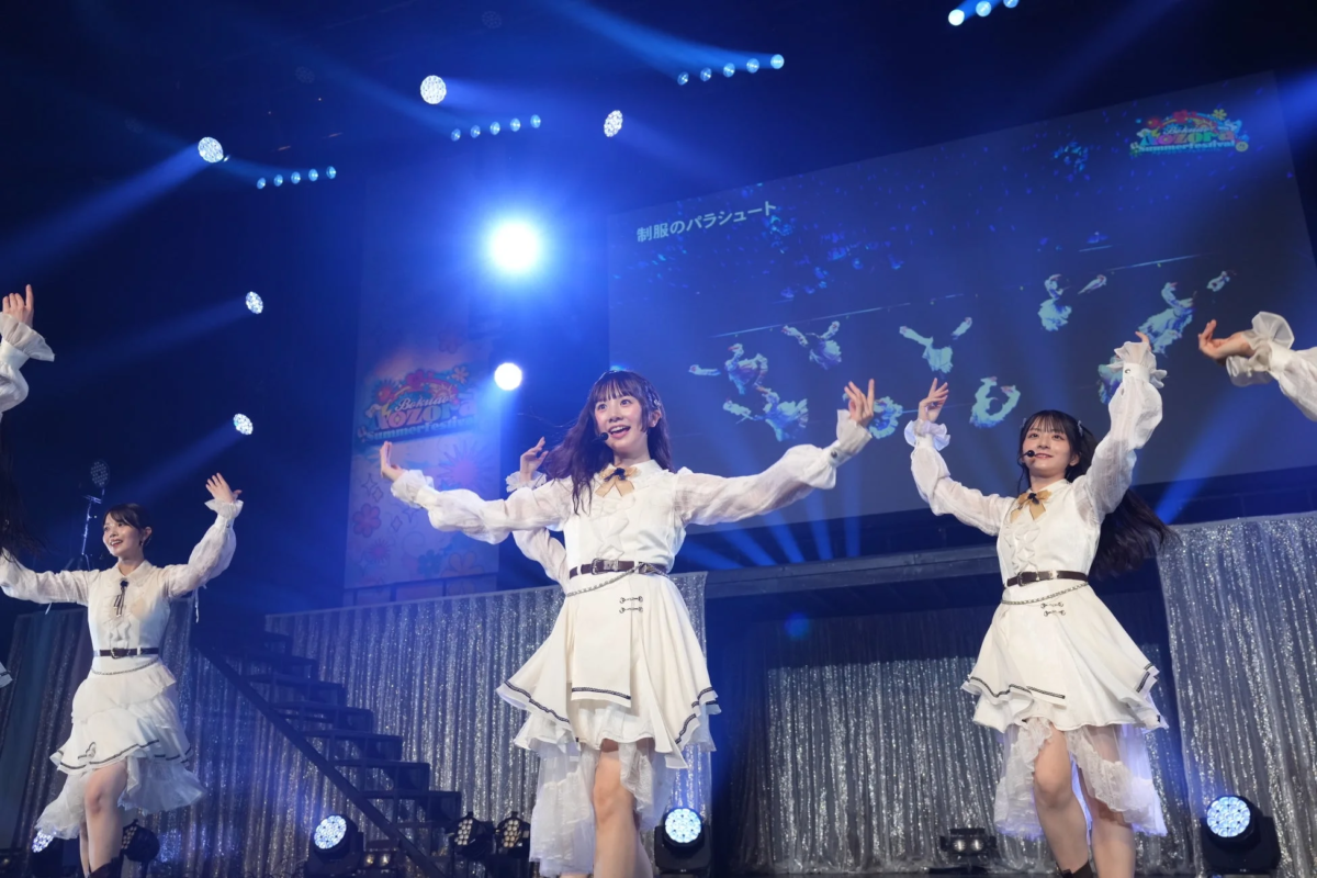 白いフリル付きの衣装をまとった複数の女性アイドルが、明るいスポットライトが当たるステージでパフォーマンスをしている様子