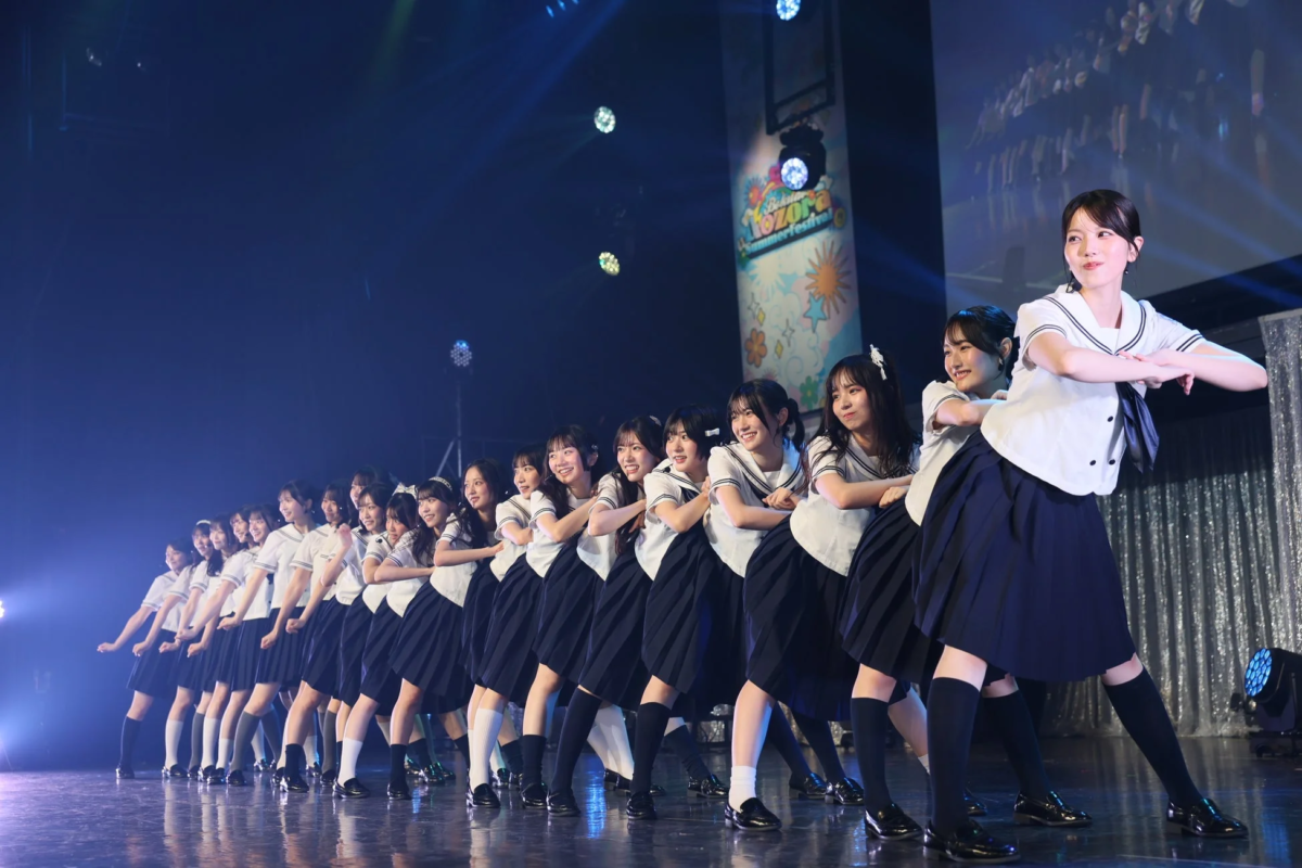 セーラー服を着た多数の女性がステージ上で一列に並び、笑顔でパフォーマンスを行っている