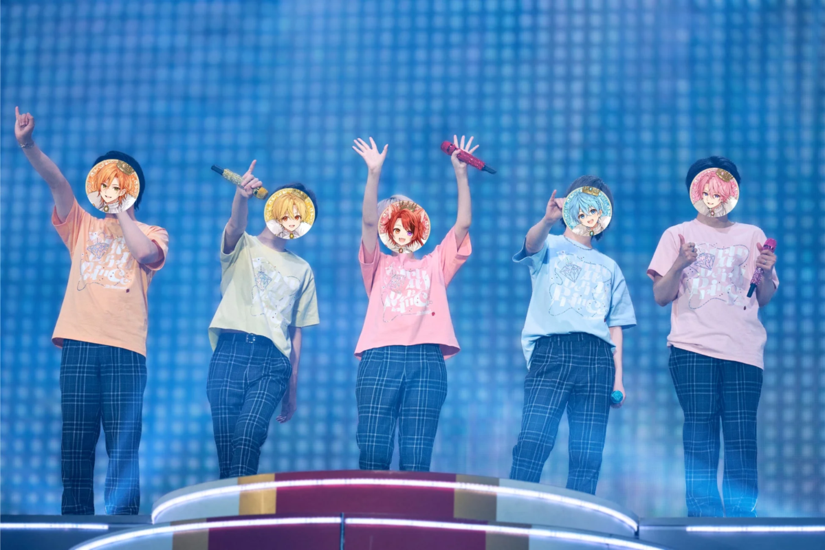 5人組のアイドルグループがステージ上でパフォーマンスをしている様子です。全員がお揃いのTシャツとチェック柄のパンツを着用し、マイクを手に歌っています。背景は青い照明で彩られています。