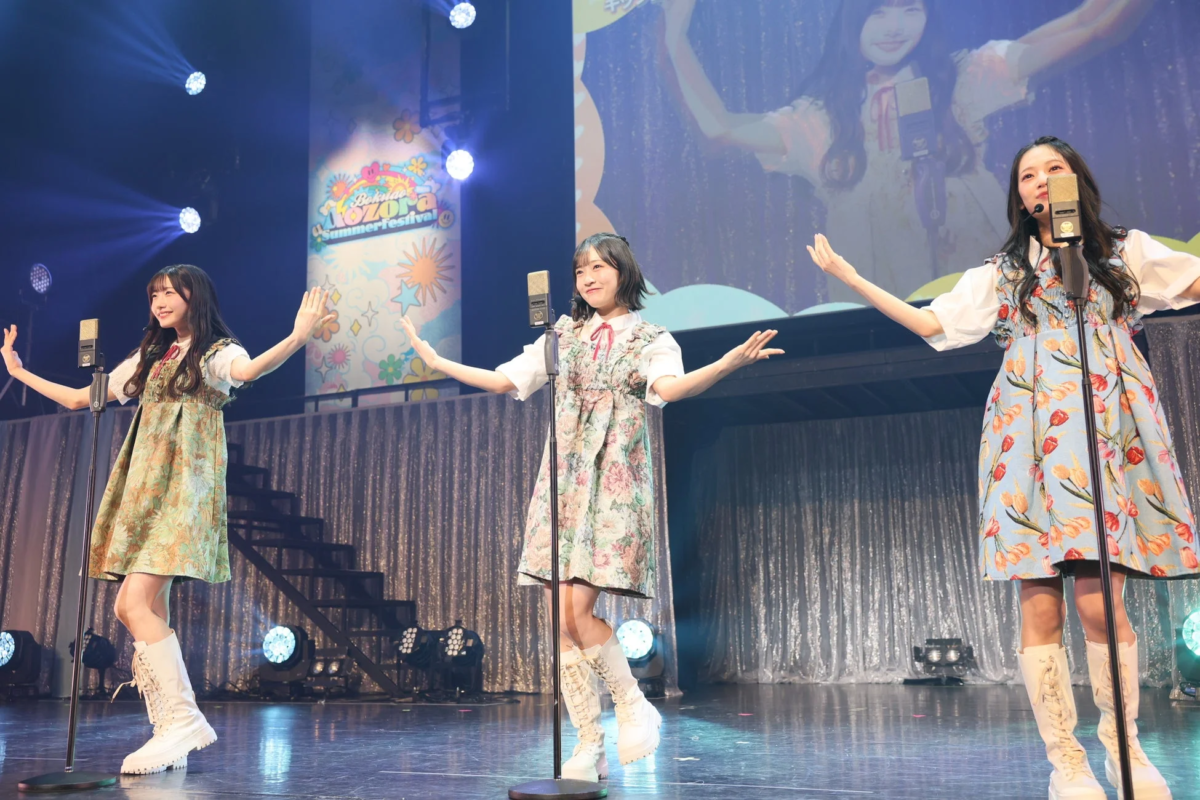 3人の女性アイドルが花柄の衣装と白いブーツを身につけ、ステージ上で笑顔でパフォーマンスしています