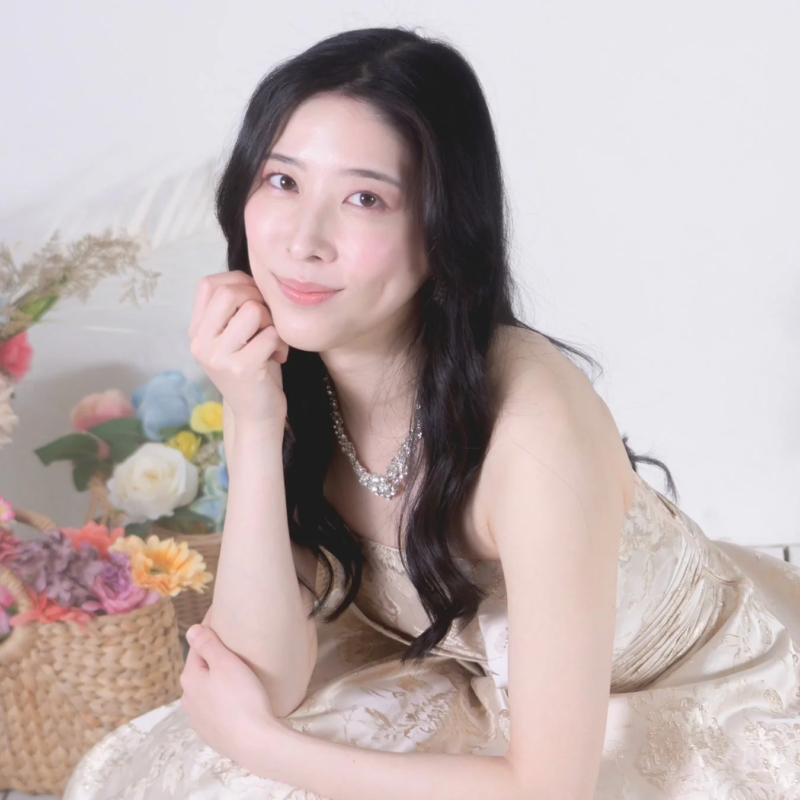 豪華なドレスとネックレスを身につけた美しい女性が、花飾りの前で優しく微笑んでいるポートレート写真。エレガントな雰囲気が魅力です。
