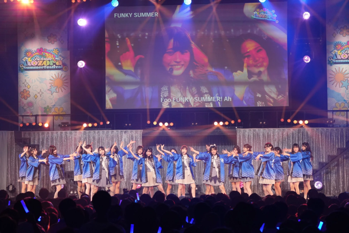 ステージで青い衣装を着た多数の女性アイドルグループがパフォーマンスを行っており、背後のスクリーンにはメンバーのアップや歌詞が映し出されています