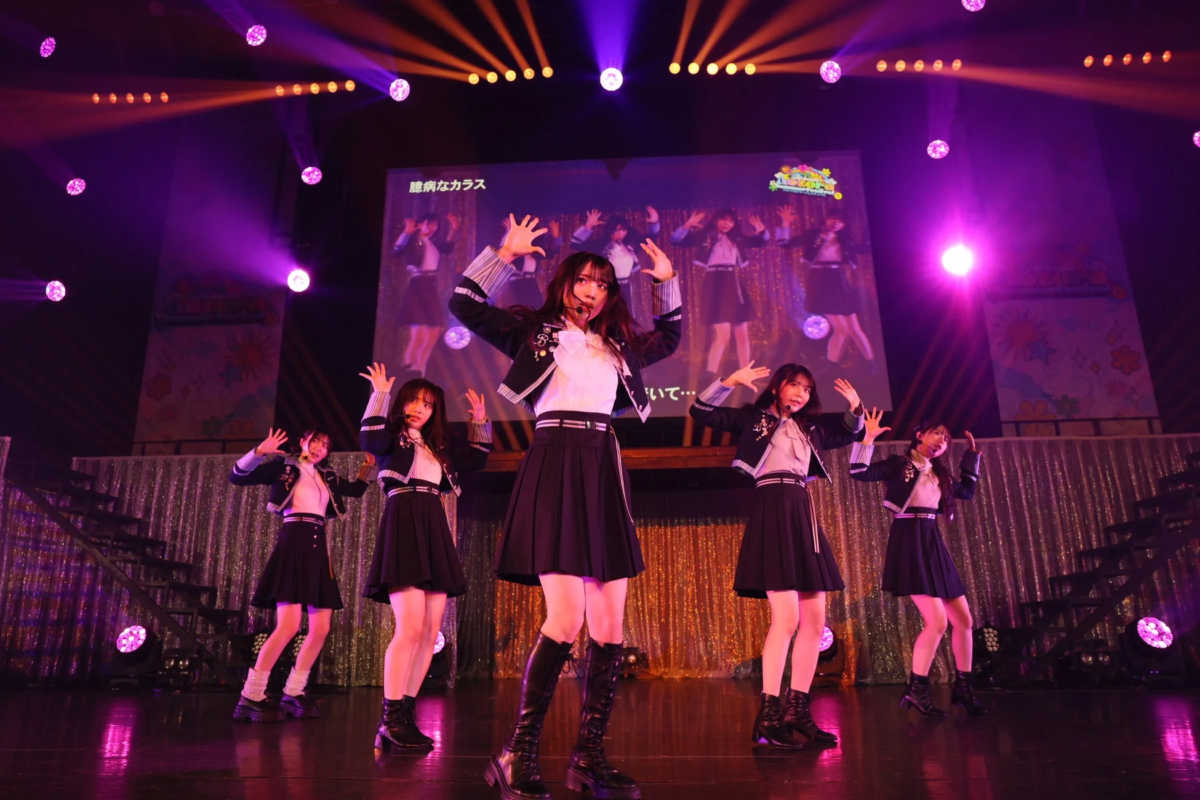 ライブステージで、制服風の衣装を着た5人の女性アイドルグループが、鮮やかな紫とオレンジの照明の下でパフォーマンスをしている様子