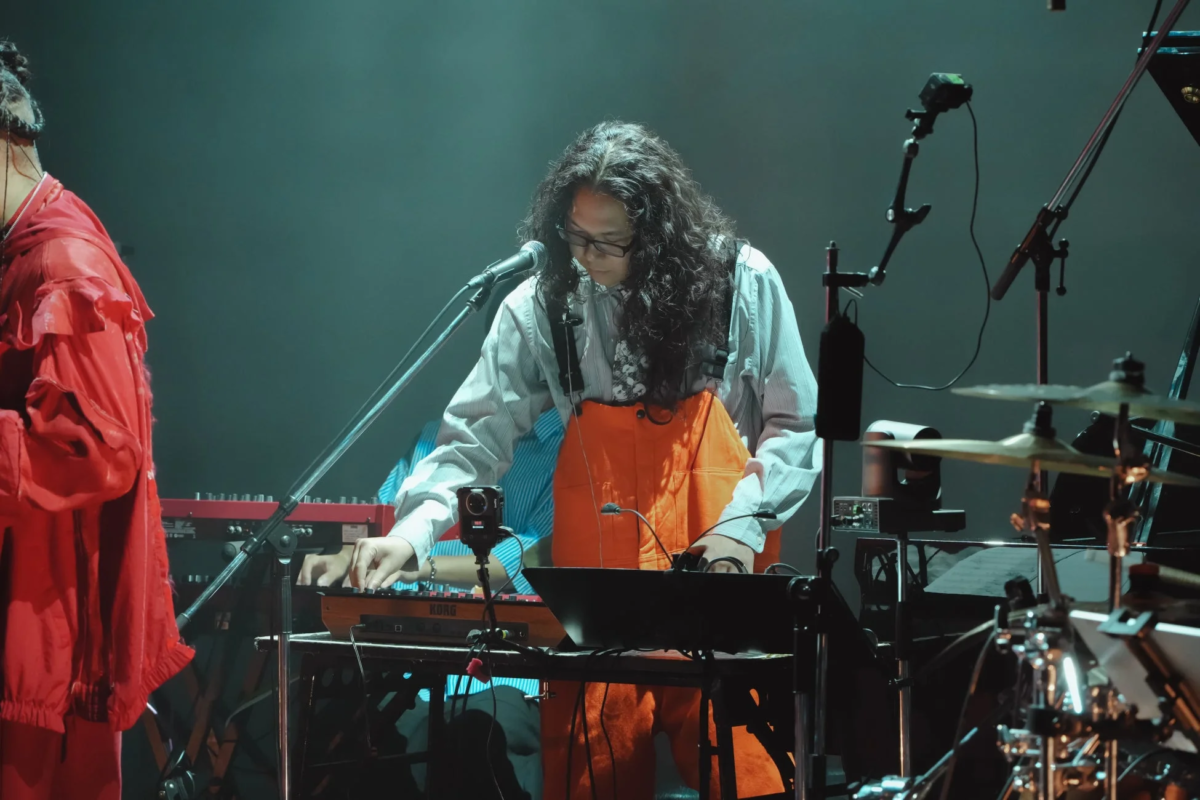 長髪と眼鏡のミュージシャンが、ライブステージでKORGのキーボードを演奏している様子