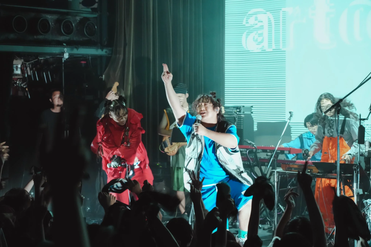 ライブステージでバンドが熱演しており、中央のボーカルがマイクを手に観客を煽っている